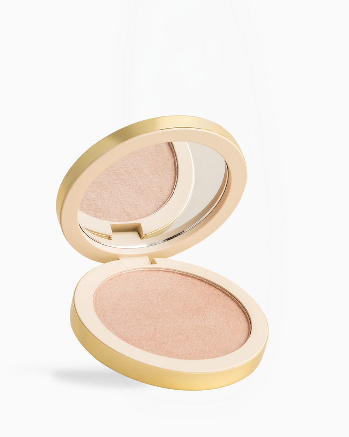 An open round makeup compact with a gold-tone case, showing a shimmering light peach-beige pressed powder and a mirror on the inside of the lid, set against a clean white background.
